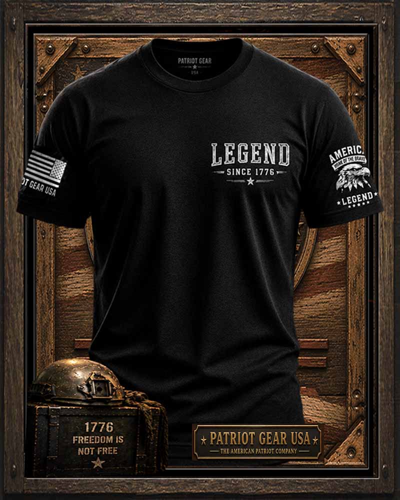A 'Patriot Gear USA' black short-sleeve graphic T-shirt displayed on a wooden mannequin torso with patriotic-themed props. The shirt has white text reading 'LEGEND SINCE 1776' on the chest, a distressed American flag graphic on the right sleeve, and a white eagle-and-text graphic on the left sleeve.