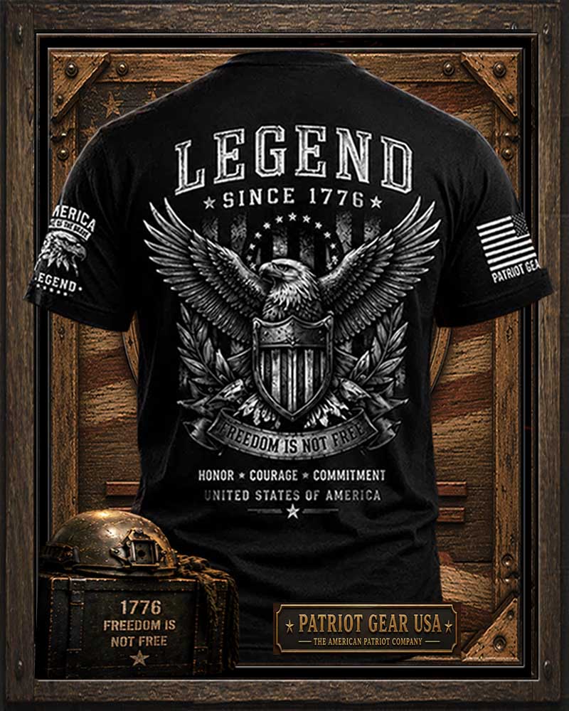 Black short-sleeve graphic T-shirt shown from the back, featuring a large white eagle and shield design with the words 'LEGEND SINCE 1776' and 'FREEDOM IS NOT FREE.' The shirt is displayed inside a rustic wooden framed graphic with 'Patriot Gear USA' branding and decorative text.