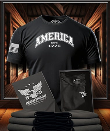 Patriot Gear USA Apparel Company Black t-shirt with 'America Est. 1776' text, displayed with folded shirts and branded American Outfitters logo on a wooden display background.
