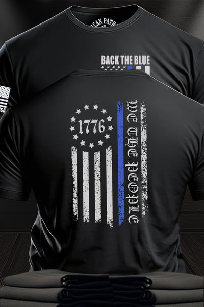 Patriot Gear USA Apparel Black t-shirt with text "Back the Blue" and a flag design with text "We the People", displayed against a wooden  American Outfitters Apparel Company background.